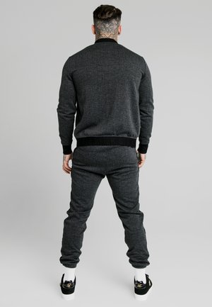 Mężczyzna z tatuażem na szyi, ubrany w ciemnoszarą, kraciastą marynarkę i dopasowane spodnie, stojący tyłem na jednolitym tle.