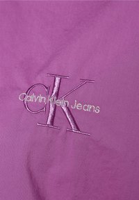Violetti kangas, jossa hopealangalla kirjailtu "CK" ja "Calvin Klein Jeans". Sileä pinta ja yksinkertainen muotoilu ilman lisäkoristeita.