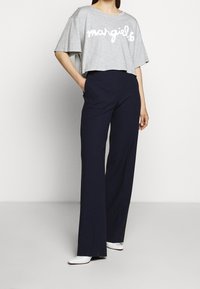 Modell i gråkortad t-shirt med texten "margiela" och marinblå vida byxor, står med ena handen i fickan. Modernt, stiligt plagg.