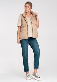 Beżowa kamizelka puchowa z wysokim kołnierzem i bocznymi kieszeniami, noszona na kremowym swetrze z dzianiny i niebieskich dżinsach. Obuwie w wzór lamparta.