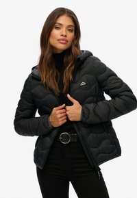 Vrouw in een zwarte gewatteerde jas met capuchon, zwarte coltrui en zwarte broek, die de rits van de jas met beide handen vasthoudt.