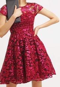 Robe en dentelle rouge avec des manches courtes, des motifs floraux, des accents de sequins, et un corsage ajusté qui se transforme en une jupe évasée. Pochette noire en main.