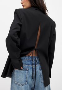 Schwarze Blazer mit offener Rückseite, an der Taille gebunden. Merkmale sind strukturierte Schultern, lange Ärmel und glattes Material, kombiniert mit blauen Denim-Shorts.