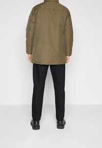 Khaki pufferjakke med lynlås, har en høj krave, lange ærmer og subtil branding på ryggen; kombineret med sorte bukser og støvler.