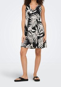 Femme portant une robe noire sans manches ornée de motifs de feuilles tropicales blanches et rose clair, assortie à des sandales à plateforme noires.