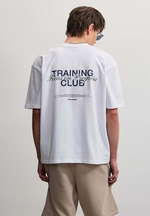Persona con el pelo corto castaño, gafas de sol, camiseta blanca con el texto "Training Focus on Progress Club" y pantalones cortos beige, vista desde atrás.