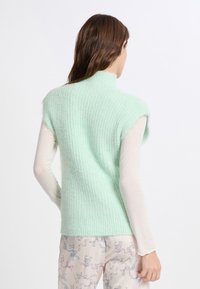Maglione senza maniche di colore verde menta con colletto alto, caratterizzato da una morbida trama e un design a coste. Indossato sopra una maglietta a maniche lunghe bianca.