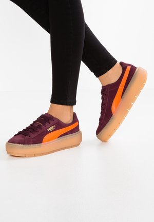 Jambes portant un pantalon noir et des baskets en daim violet avec une bande orange et des semelles épaisses en caoutchouc translucide sur un sol blanc.