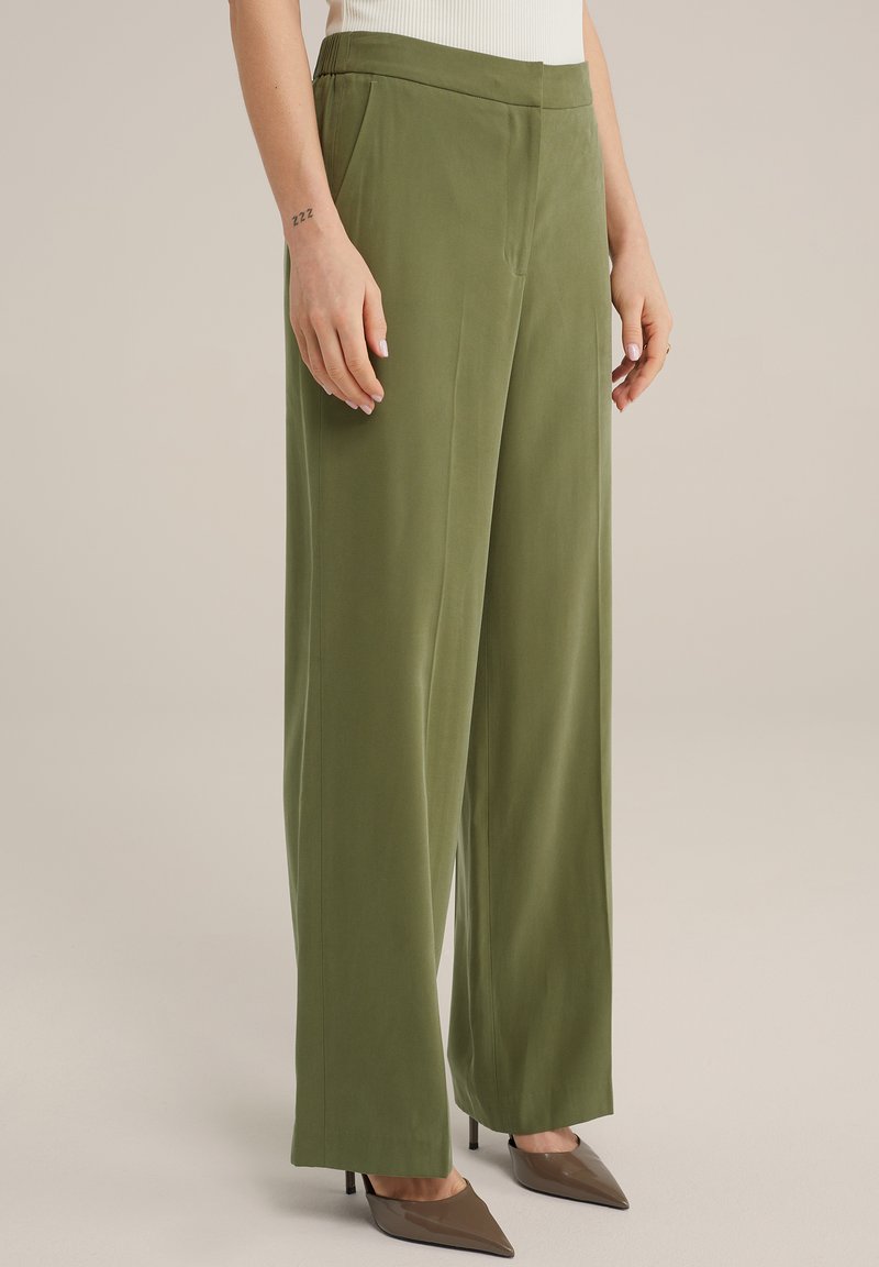 Pantalones verdes de pierna ancha, confeccionados en una tela suave con cintura alta, que presentan bolsillos laterales y un ligero brillo, combinados con unos zapatos marrones de punta.
