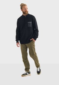 Schwarzer Reißverschluss-Pullover mit strukturiertem Strick, ausgestattet mit einer einfarbigen schwarzen Brusttasche. Kombiniert mit olivgrünen Hosen und schwarzen Sneakers mit weißen Akzenten.