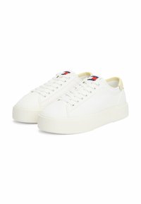 Baskets en toile blanches avec une semelle épaisse et texturée, dotées d'un bout rond, d'un système de laçage et d'une étiquette logo rouge, blanc et bleu sur la languette.
