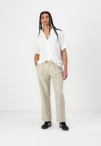 Camicia bianca a maniche corte con bottoni e un motivo testurizzato, abbinata a pantaloni beige chiaro e sneakers nere con strisce bianche.