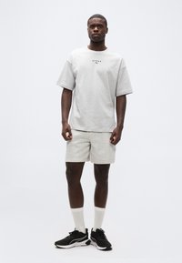 Άνδρας όρθιος, φορώντας oversized ανοιχτό γκρι μπλουζάκι με λογότυπο "HYROX", ασορτί σορτσάκι, λευκές κάλτσες και μαύρα αθλητικά παπούτσια, σε απλό λευκό φόντο.