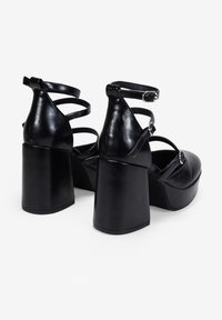 Zapatos de plataforma negros con acabado de cuero brillante, tacones anchos y múltiples correas delgadas en los tobillos con detalles de hebilla.