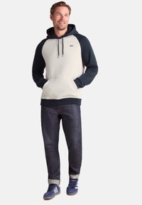 Light gray and navy blue hoodie with a front pocket, drawstring hood, ribbed cuffs, and contrasting raglan sleeves. Worn with dark jeans and blue sneakers.