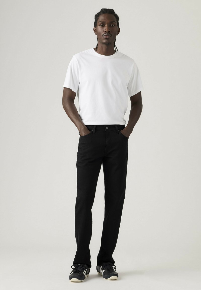 Mann steht mit den Händen in den Taschen, trägt ein einfaches weißes T-Shirt, schwarze Jeans und schwarze Sneakers vor einem grauen Hintergrund.