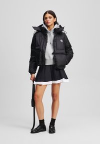 Doudoune noire courte avec capuche, sweat à capuche gris en dessous, jupe plissée noire avec garniture blanche, chaussures noires et sac à bandoulière.