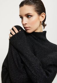 Crna pulover s visokim ovratnikom, mekane teksture i metalik sjajem, s dugim, prevelikim rukavima koji prelaze preko ruku.