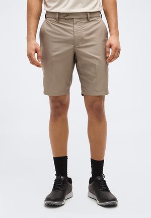 Mężczyzna w beżowych, dopasowanych szortach, czarnych skarpetkach i czarnych trampkach, stojący na jasnym, jednolitym tle.