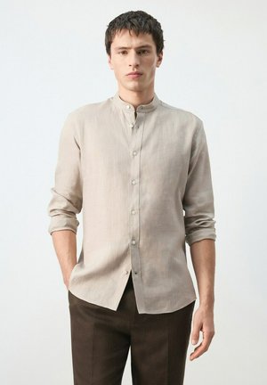 Homme portant une chemise beige à col mandarin avec les manches retroussées et un pantalon marron foncé, debout devant un fond clair uni.