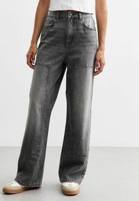 Vaqueros de pierna ancha en denim gris deslavado, con grandes bolsillos tipo parche y costuras en contraste. Llevados con zapatillas blancas y beige.