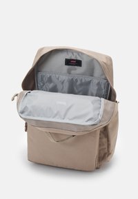 Sac à dos beige avec un intérieur gris. Il possède une fermeture à rabat et un compartiment zippé. Fabriqué dans un tissu durable avec une texture douce.