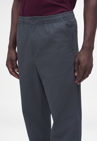 Gros plan d'une personne portant un pantalon gris foncé à taille élastique avec des poches latérales et une chemise bordeaux rentrée.