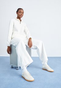 Witte fleece pullover met een ritskraag, brede witte broek en beige sneakers met rubberen zolen. Model zittend op een metalen container.