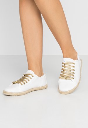 Vita sneakers med guldmetalliska snören, rund tå och jute-utförande runt sulan. Slät textur, avslappnad design och lågprofil.