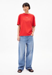 Rotes Baumwoll-T-Shirt mit kurzen Ärmeln und Rundhalsausschnitt, kombiniert mit hellblauen weiten Jeans und schwarzen Sandalen. Einfaches Logo auf der Brust.