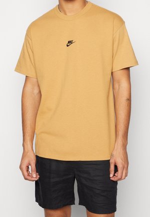 Person trägt ein senfgelbes Nike-T-Shirt mit schwarzem Logo in der Mitte der Brust und schwarze Shorts, steht mit entspannten Armen.