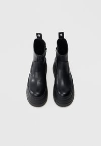 Svarte ankelstøvler i skinn med en kraftig yttersåle, sideskjermer og preget merke på sålen. Jevn tekstur med et minimalistisk design.