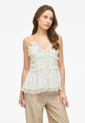 Młoda kobieta z długimi brązowymi włosami ubrana w białą, bezrękawnikową bluzkę z falbankami i beżowe spodnie na jednolitym tle.