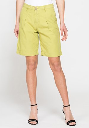 Donna con pantaloncini verde chiaro fino al ginocchio e sandali neri con tacco alto, in piedi davanti a uno sfondo semplice.