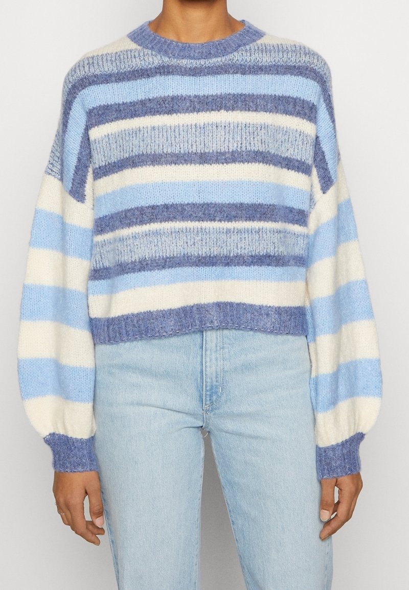Personne portant un pull doux rayé aux tons bleu, blanc et violet, associé à un jean bleu clair, debout devant un fond uni.