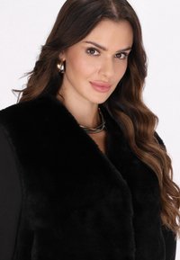Vesta din blană artificială neagră cu guler înalt, având o textură moale. O colier argintie este purtat, iar fundalul este neutru.