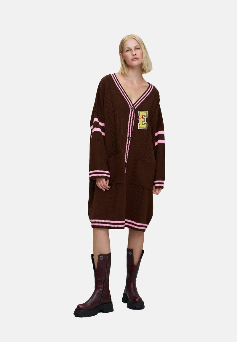 Femme blonde portant un long cardigan marron avec des rayures roses et un écusson "E", associé à des bottes marron foncé montant jusqu'aux genoux.