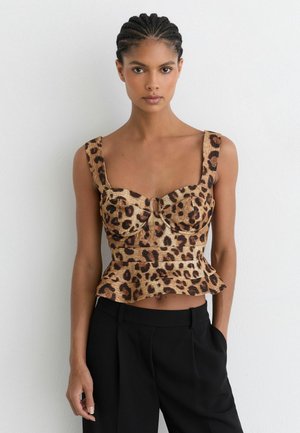 Frau mit geflochtenem Haar trägt ein ärmelloses Oberteil mit Leopardenmuster und Peplum-Saum sowie schwarze Hosen, steht vor einer weißen Wand.