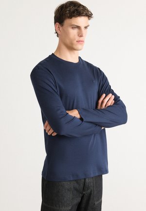 Young man with folded arms wearing a navy blue long-sleeve shirt and dark jeans against a plain light background.