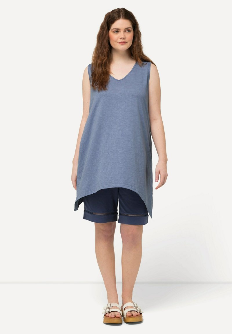 Túnica azul sin mangas con escote en V, que presenta un dobladillo asimétrico; combinada con pantalones cortos azul marino con acentos de malla y sandalias blancas.