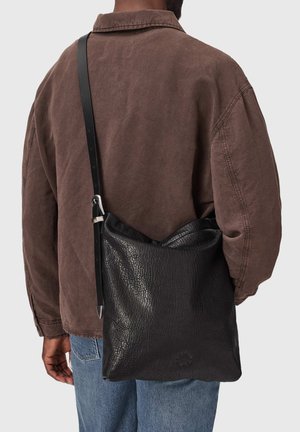 Mand iført brun jakke og blå jeans med en sort tekstureret lædertaske over skulderen, vist bagfra.