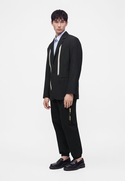 Homme portant une veste de costume noire avec un détail de ruban à mesurer, pantalon assorti avec poche zippée, chemise bleu clair et mocassins noirs, debout devant un fond blanc.