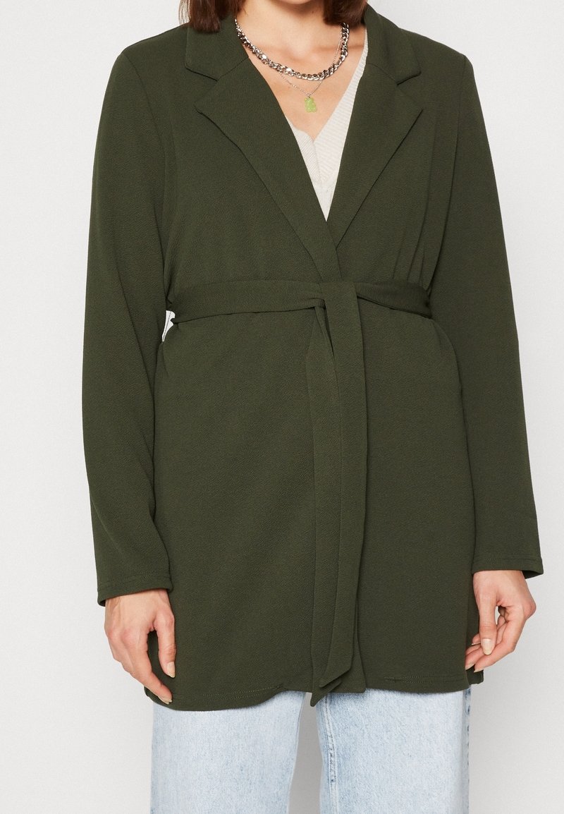 Blazer largo verde con textura, con cintura ceñida y apertura frontal, que presenta un cuello con muescas y mangas largas, combinado con jeans azul claro.