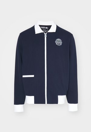 Marinblå zip-jacka gjord av texturerat tyg, med vita accenter, krage och en broderad tennis klubb-logotyp på vänster bröst.