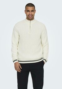 Suéter de punto blanco con cuello alto y cierre de media cremallera. Presenta rayas en blanco y negro en los puños y el dobladillo.