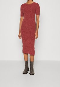 Rotes geripptes Strickkleid mit Rundhalsausschnitt und kurzen Ärmeln, mit seitlichen Schlitzen und kombiniert mit grauen Knöchelstiefeln. Glatte Textur.
