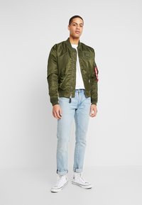 Olivgrüne Bomberjacke mit einem Reißverschluss auf der Vorderseite, elastischen Bündchen und Patch-Details, getragen über einem weißen T-Shirt und hellblauen Jeans.