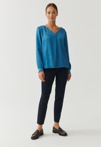 Blusa blu in seta con scollo a V e maniche lunghe arricciate, abbinata a pantaloni aderenti blu scuro e mocassini marroni su uno sfondo neutro.