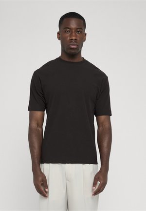 Sort kortærmet T-shirt med rund hals, båret af en stående mand med lyse beige plisserede bukser mod en ensfarvet grå baggrund.