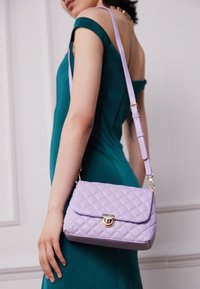 Borsa a mano trapuntata lavanda con chiusura dorata e tracolla regolabile. Presenta una forma rettangolare con una texture liscia e una leggera lucentezza.